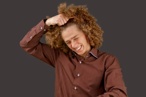灰色の背景に茶色のシャツを着た長髪の巻き毛の男は 木製の櫛を使用しています 美容室で散髪する前の感情 痛みを感じる — ストック写真