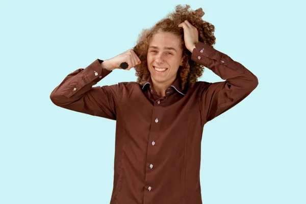 青い背景の茶色のシャツを着た長髪の巻き毛の男は 木製の櫛を使用しています 美容室で散髪する前の感情 痛みを感じる — ストック写真