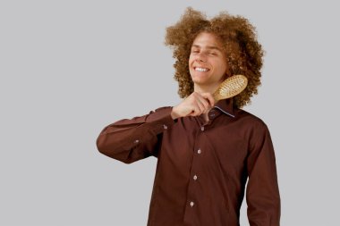 Gri arka planda kahverengi gömlekli, uzun saçlı, kıvırcık saçlı bir adam ahşap tarak kullanıyor. Bir kuaförde saç tıraşından önce duygular.