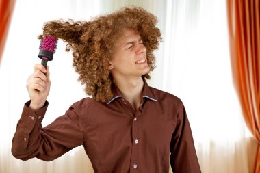 Uzun saçlı, kahverengi gömlekli kıvırcık saçlı bir adam metal yuvarlak tarak kullanıyor. Bir kuaförde saç tıraşından önce duygular. Taramaktan dolayı acı.