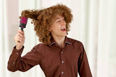 Uzun saçlı, kahverengi gömlekli kıvırcık saçlı bir adam metal yuvarlak tarak kullanıyor. Bir kuaförde saç tıraşından önce duygular. Taramaktan dolayı acı.