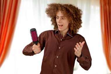 Uzun saçlı, kahverengi gömlekli kıvırcık saçlı bir adam metal yuvarlak tarak kullanıyor. Bir kuaförde saç tıraşından önce duygular.