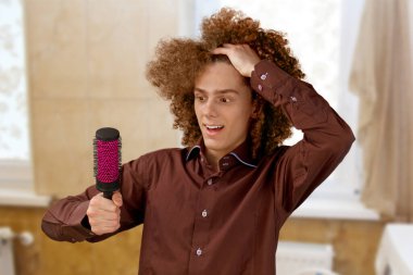 Uzun saçlı, kahverengi gömlekli kıvırcık saçlı bir adam metal yuvarlak tarak kullanıyor. Bir kuaförde saç tıraşından önce duygular.