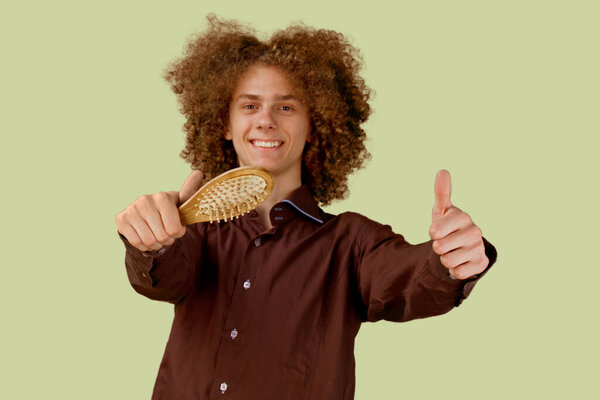 A long-haired curly-haired guy in a brown shirt on a beige background uses a wooden comb. Emotions before a haircut in a hairdresser.