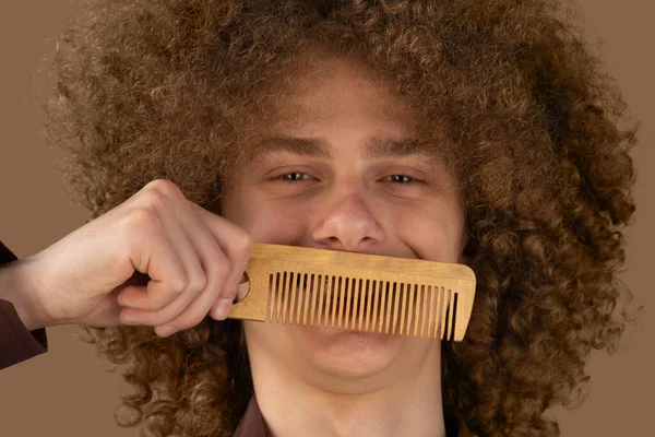 茶色の背景に茶色のシャツを着た長髪の巻き毛の男は 木製の櫛を使用しています 美容室で散髪する前の感情 — ストック写真