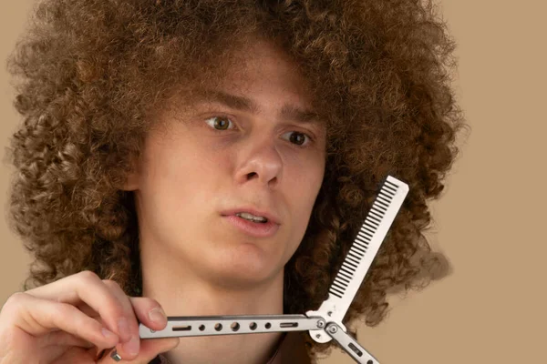 A long-haired curly-haired guy in a brown shirt on a brown background uses a metal comb. Emotions before a haircut in a hairdresser.