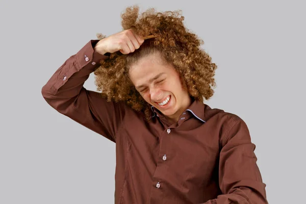 灰色の背景に茶色のシャツを着た長髪の巻き毛の男は 木製の櫛を使用しています 美容室で散髪する前の感情 痛みを感じる — ストック写真