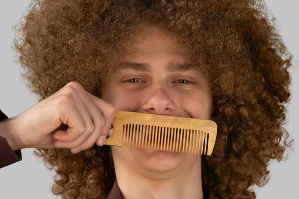 灰色の背景に茶色のシャツを着た長髪の巻き毛の男は 木製の櫛を使用しています 美容室で散髪する前の感情 — ストック写真