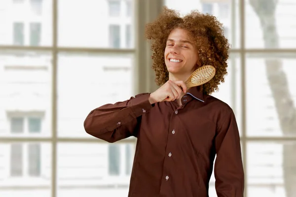 A long-haired curly-haired guy in a brown shirtuses a wooden comb. Emotions before a haircut in a hairdresser.