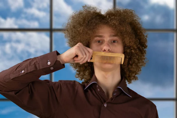 A long-haired curly-haired guy in a brown shirtuses a wooden comb. Emotions before a haircut in a hairdresser.