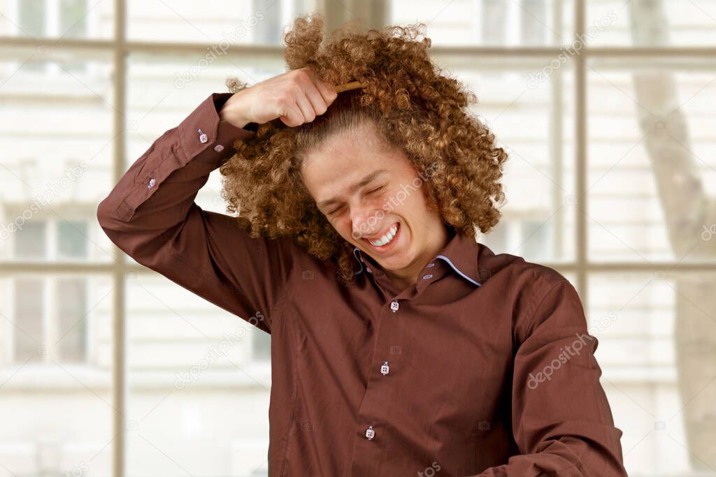 A long-haired curly-haired guy in a brown shirt uses a wooden comb. Emotions before a haircut in a hairdresser. Pain from combing.