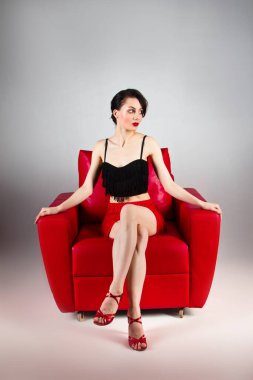 A beautiful brunette in red shoes sits in a red armchair on a gray background. Latin dances. The color of passion