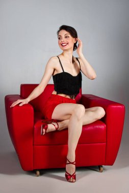 A beautiful brunette in red shoes sits in a red armchair on a gray background. Latin dances. The color of passion