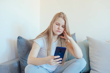 Sıkılmış genç bir kız akıllı bir telefon kullanıyor ve oturma odasında gri bir kanepede oturuyor.