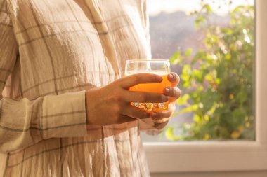 Genç bir kadın taze ve lezzetli sıkılmış portakal suyu içer, sağlıklı ve vitamin doludur, sabahları, evde, kahvaltı etmeden ve pencerenin kenarında güneşin tadını çıkarmadan önce..