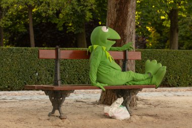 Madrid, İspanya - 20 Haziran 2020: Kurbağa Kermit kılığına girmiş bir erkek ya da kadın, geçimini sağlamak için Madrid, Retiro Park 'taki ahşap bir bankta dinleniyor..