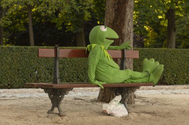 Madrid, Spain - June 20, 2020: A man or woman, disguised as Kermit the frog, to earn a living, sits resting on a wooden bench in Retiro Park, Madrid. clipart