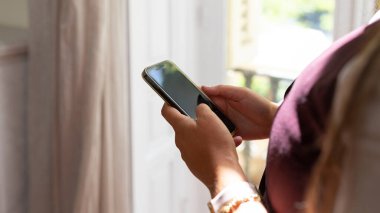 Bir kişi parlak ışığı olan bir odanın içinde duruyor ve akıllı telefona bakıyor. Odaklandıkları şey onların elleri ve cihaz. Ortalık sakin ve rahat..