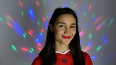 Young attractive woman in a new year costume smiles broadly against the background of a multi-colored garland. Close-up portrait of a young happy woman, she in a festive mood tries on a santa claus costume, preparing for the new year.