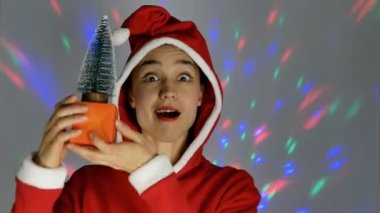 Noel Baba gibi giyinmiş güzel genç bir kadın gülümsüyor ve elinde küçük bir Noel ağacı tutuyor. Yeni yıl tatili.