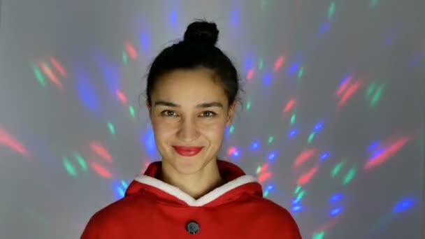 Close-up portret van een jonge gelukkige vrouw, in een feestelijke stemming droomt ze van nieuwjaarsgeschenken, glimlacht en zwaait met haar hand in begroeting, bereidt zich voor op Kerstmis. — Stockvideo