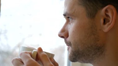 Mutfakta dikilmiş beyaz bir fincandan kahve içen genç bir adamın yakın çekimi. Genç ve çekici bir adam beyaz bir fincandan kahve içip pencereden dışarı bakıyor..