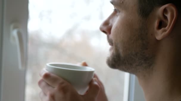 一个年轻人站在厨房里用白杯喝咖啡的特写镜头。一个年轻迷人的男人用白杯喝咖啡，看着窗外. — 图库视频影像