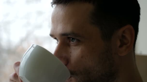 Großaufnahme eines jungen Mannes, der in seiner Küche steht und Kaffee aus einer weißen Tasse trinkt. Ein junger attraktiver Mann trinkt Kaffee aus einer weißen Tasse und schaut aus dem Fenster. — Stockvideo