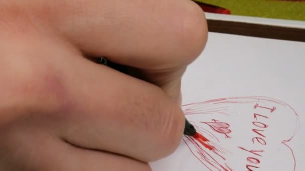 Молодой парень рисует валентинку для своей девушки. Крупным планом руки молодого человека он пишет на бумаге слова, которые я люблю тебя красной ручкой. — стоковое видео