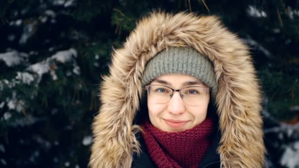 Mujer atractiva con un sombrero verde y capucha esponjosa camina en un parque cubierto de nieve. Joven mujer atractiva sonriendo y mirando a la cámara en el fondo del bosque de invierno. Joven mujer feliz en un — Vídeos de Stock