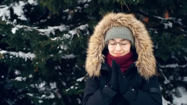 Joven mujer feliz en un ambiente romántico se calienta las manos mientras camina en el bosque de invierno. Atractiva mujer con sombrero verde y capucha esponjosa camina en un parque cubierto de nieve. — Vídeo de stock