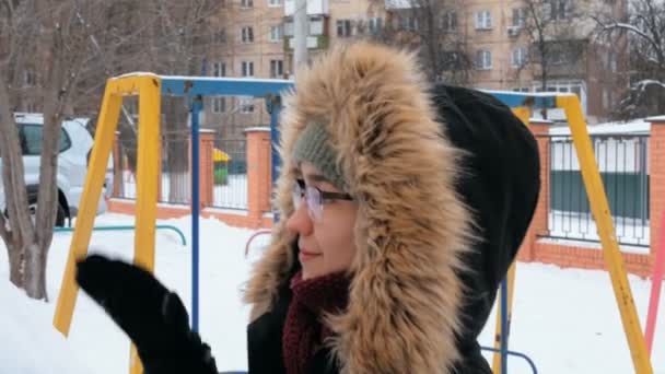 Una giovane donna sorridente scolpisce un pupazzo di neve nel suo giardino. Una donna attraente con un cappello verde e un cappuccio soffice mette una carota facendo un naso per un pupazzo di neve. Giovane donna felice in uno stato d'animo romantico scolpisce un — Video Stock