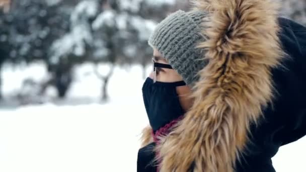 Une jolie femme coiffée d'un chapeau vert et d'une capuche moelleuse, respectant les règles de la protection individuelle, se promène dans un parc enneigé. Une jeune femme séduisante dans un masque de protection et des lunettes contre le — Video