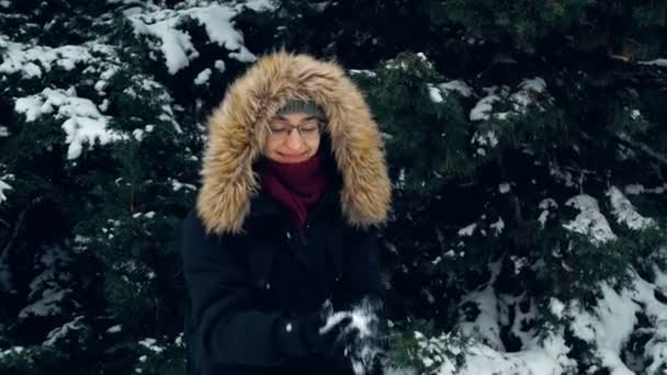 Uma mulher atraente com um chapéu verde e um capuz fofo caminha em um parque coberto de neve, joga neve com as mãos e é genuinamente feliz. Jovem mulher atraente joga neve sobre si mesma e alegremente bate palmas — Vídeo de Stock
