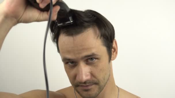 Um jovem cara engraçado corta o cabelo usando um cortador de cabelo enquanto está sentado em casa em quarentena. Um jovem alegre corta seu próprio cabelo com um aparador elétrico. — Vídeo de Stock