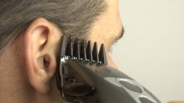 Genç ve çekici bir adam karantinada evde otururken saç makasıyla saçını kesiyor. Genç bir adam elektrikli makasla kendi saçını kesiyor..