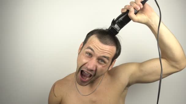Der Mann rasiert sich den Kopf und schreit laut in den Spiegel. Ein junger lustiger Kerl vor dem Spiegel rasiert sich die Haare mit einem Haarschneider, während er zu Hause in Quarantäne sitzt. — Stockvideo