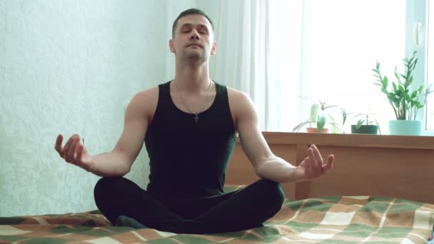 若い幸せな男は自宅でベッドの上にハスの位置に座って瞑想に従事しています。若い男が自宅で瞑想を楽しんでいる。若い魅力的な男は彼のリビングルームのベッドで瞑想しています. — ストック動画