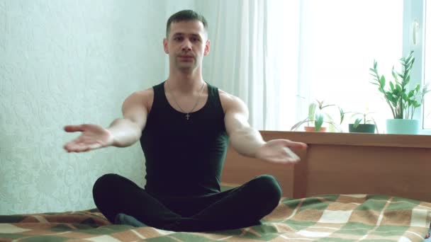 Fiatal, vonzó férfi, aki assanákat ad elő az ágyon ülve a nappalijában. Fiatal srác légzési gyakorlatokat végez és otthon meditál. Fiatal boldog ember meditál, miközben lótuszban ül — Stock videók