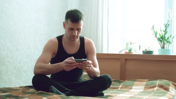 Giovane uomo felice scorrendo i social media feed sul suo smartphone mentre seduto sul letto a casa. Giovane uomo attraente scrivere e-mail sul suo smartphone mentre seduto sul letto nel suo soggiorno — Video Stock