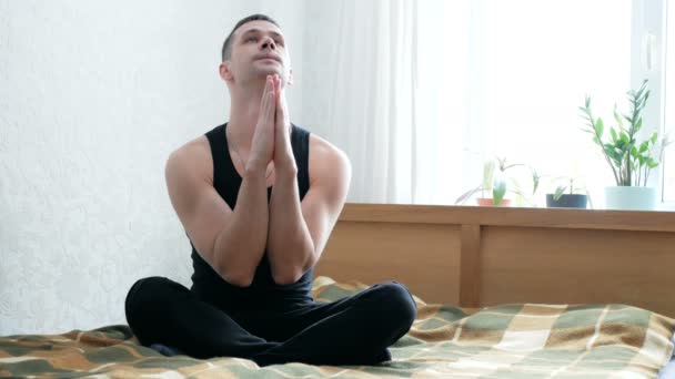 Joven hombre atractivo realizando assanas sentado en la cama en su sala de estar. Joven hombre feliz haciendo meditación mientras está sentado en posición de loto en la cama en casa. Joven chico haciendo ejercicios de respiración — Vídeos de Stock