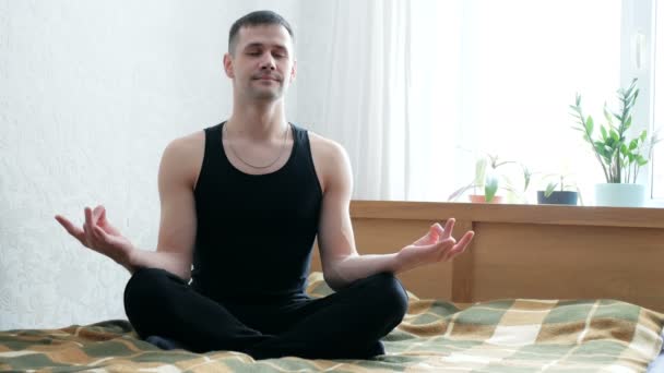 Joven hombre feliz se dedica a la meditación sentado en posición de loto en la cama en casa. Joven atractivo hombre está meditando en la cama en su sala de estar.Joven chico está disfrutando de la meditación en casa. — Vídeos de Stock