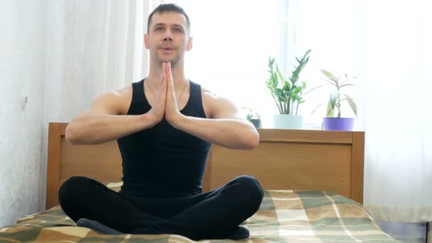 Joven hombre atractivo realizando assanas sentado en la cama en su sala de estar. Joven hombre feliz haciendo meditación mientras está sentado en posición de loto en la cama en casa. Joven chico haciendo ejercicios de respiración — Vídeos de Stock