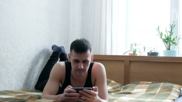 Jeune homme attrayant utilise smartphone assis sur le canapé. Jeune homme tapant un message sur son smartphone tout en étant assis sur le lit à la maison. — Video