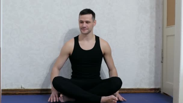 Joven hombre feliz haciendo meditación sentado en posición de loto en la esterilla de yoga en casa. El joven está haciendo yoga y meditando en casa. Concepto de estilo de vida saludable. Joven atractivo hombre realiza assanas — Vídeos de Stock