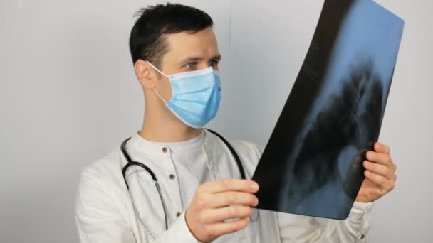 Ein junger, gut aussehender Arzt in medizinischer Maske hält ein Röntgenbild der Lungen in den Händen und führt eine Analyse durch. Ein junger Chirurg mit medizinischer Schutzmaske untersucht ein Röntgenbild eines Patienten — Stockvideo