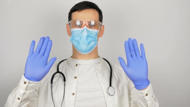 El joven médico agita su mano amablemente, dando la bienvenida al paciente.. Joven médico guapo se pone gafas de seguridad y una máscara de protección médica mientras se prepara para examinar a los pacientes. — Vídeos de Stock