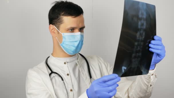 Mladý chirurg v ochranné lékařské masce vyšetřuje rentgen plic pacienta a zvažuje diagnózu. Mladý pohledný doktor v lékařské masce drží rentgenový snímek plic v jeho — Stock video