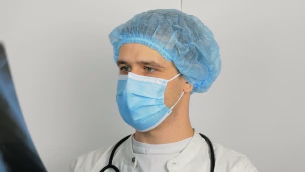 Ein junger Chirurg mit medizinischer Schutzmaske untersucht ein Röntgenbild der Lunge eines Patienten und erwägt die Diagnose. Ein junger, gut aussehender Arzt in medizinischer Maske hält ein Röntgenbild der Lungen in der Hand. — Stockvideo
