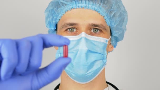 Un beau jeune médecin examine un nouveau médicament. Un jeune beau médecin dans un masque médical tient une pilule rouge dans ses mains présentant un nouveau médicament. — Video
